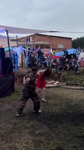 Two people are dancing outdoors in front of an audience. The person on the left is wearing a camouflage outfit, and the person on the right is wearing a red dress. They are holding hands and appear to be dancing a waltz or similar partner dance. The audience is sitting and standing on a grassy slope behind the dancers. In the background, there are tents, a building with graffiti, and a sign that says "ИНФО." The lighting suggests it is either early morning or late evening.
