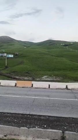 The video pans across a rural landscape with rolling green hills under a cloudy sky. In the foreground, there is a paved road with white and orange concrete barriers along its edge. Further back, scattered houses and structures are visible on the hillsides. The overall impression is one of a remote, possibly mountainous, rural setting.