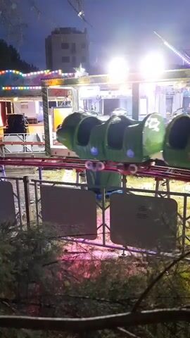 The video shows a small, green, caterpillar-shaped roller coaster at an amusement park at dusk. The roller coaster is moving along its tracks, with one car visible in the foreground carrying a person. In the background, there are bright lights and other amusement park rides, including a building structure. The area is partially obscured by trees and bushes in the foreground. The atmosphere appears to be that of a lively evening at an amusement park.