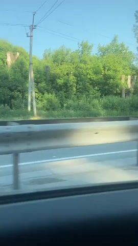 The video captures a view from inside a moving car. The passenger's side door panel, interior door handle, and a side mirror are visible in the foreground. Through the side mirror and the car window, a highway with a guardrail and trees lining the road can be seen. The sky is blue and appears to be daytime.