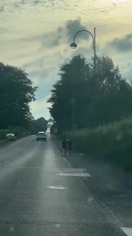 The video shows a road with two people walking on the side. The person on the left is wearing a black t-shirt and black shorts. The person on the right is wearing a black t-shirt and black shorts. There is a car driving on the road in front of them. There are trees on the side of the road and a lamppost. The sky is cloudy.