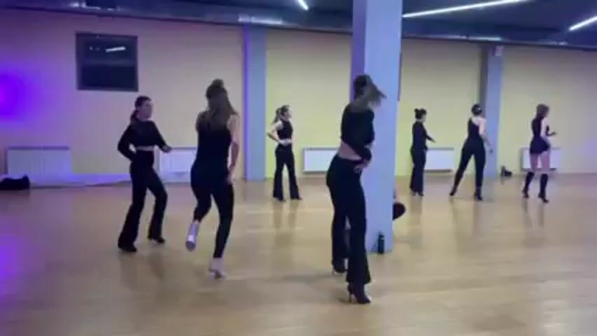 In a dance studio with a light wood floor, several women are participating in a dance class. All are dressed in black outfits. There is a white pillar in the center of the room. Several windows are visible in the background, and a white radiator is mounted on the wall. Some of the dancers are wearing heels. The dancers are practicing a choreographed routine. The instructor is in the front, giving instructions, and the other dancers are following her lead. The mood is focused and energetic.