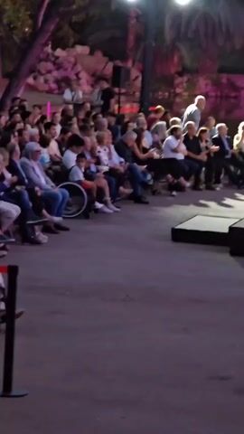 A group of people are seated in the foreground of the video, facing a stage. The audience is composed of individuals of various ages and genders. Some are casually dressed, while others are in more formal attire. Several people are holding phones and appear to be recording or taking pictures. The stage consists of a series of black platforms of varying heights. A man walks across the stage from the right side of the screen. Behind the audience, there is a body of water reflecting the lights. The background features lush foliage and trees, illuminated by spotlights and ambient lighting. The overall atmosphere suggests an outdoor event, possibly a performance or presentation.