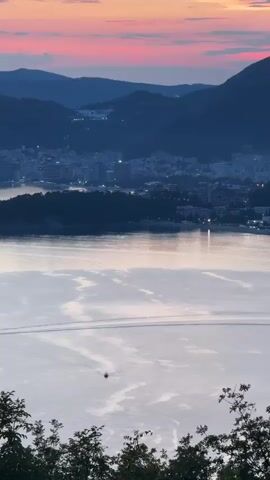 A vertical video captures a panoramic view of a coastal city at dusk. The sky is ablaze with hues of pink and orange, transitioning into a deep blue as the sun sets behind a range of dark mountains. In the foreground, lush green foliage frames the bottom of the shot. Below the mountains, a sprawling city unfolds, its buildings illuminated by the soft glow of streetlights and interior lights. The city is nestled along a curved coastline, bordering a tranquil body of water. The water reflects the vibrant colors of the sunset, creating a shimmering, almost ethereal surface. A solitary boat is visible in the distance, leaving a subtle wake as it traverses the calm sea. The overall atmosphere is one of serenity and natural beauty.