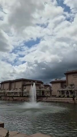 A vertical video shows a serene view of a temple complex with a large body of water and a fountain in the foreground. The architecture features ornate designs and multiple buildings. A tall, golden statue of a figure with raised arms stands prominently in the center, with smaller structures and decorative elements surrounding it. The sky is filled with dramatic, cloudy formations, suggesting a dynamic weather. Several individuals are visible in the background, walking around the temple grounds. Flowers add pops of color to the manicured landscaping. The water reflects the overcast sky, creating a peaceful ambiance.