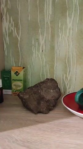 The video shows a shelf with several decorative items. On the left is a box of rosemary. Next to it is a dark, rough-textured rock. In the center, there is a green ceramic candle holder with a red saucer. To the right of the candle holder is a large, flat rock painted with a landscape scene featuring mountains, a body of water, trees, and a sun. The background is a wall with a green and white patterned wallpaper. The shelf is made of wood-grain material. The camera pans slowly from left to right, showcasing each item.