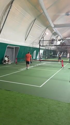 A paddle sport is being played indoors on a green court under a white tent-like ceiling. In the foreground, a man wearing an orange shirt and blue shorts is positioned on the left side of the court, holding a paddle. He is mid-swing, striking a ball over the net. Two women are on the opposite side of the net, one in a black outfit and the other in a white outfit, seemingly playing against the man and his partner, who is not fully visible. Other players and spectators are present in the background, some sitting on benches. The court has white lines marking its boundaries and a net dividing it. The environment appears to be a sports facility.