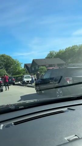 A white sedan is crashed into a dark-colored SUV on a residential street. Several people are standing around the vehicles. One person in a red shirt appears to be talking to another person near the white sedan. The sky is blue with scattered clouds, and there are trees and houses in the background. The camera is inside a vehicle, looking out through the windshield.