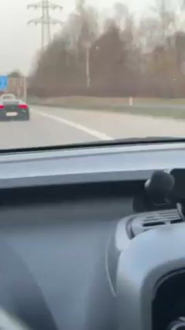 The video, shot from the inside of a moving car, shows a gray Lamborghini Huracan driving on a highway. The camera car is following the Huracan, maintaining a safe distance. The Huracan is in the left lane of the highway. There are other cars on the road. The road has multiple lanes and is bordered by trees. Overhead power lines are visible. The weather is overcast. The dashboard of the camera car is visible in the foreground. A phone holder is mounted on the dashboard. The Lamborghini has yellow brake calipers.