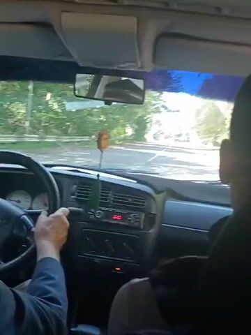 A first-person perspective video shows the interior of a car as it drives down a tree-lined road. A person's hands are visible on the steering wheel. The dashboard shows a radio and climate controls. A decorative air freshener hangs from the rearview mirror. Sunlight filters through the trees, creating dappled light on the road ahead. Another car is visible in the distance. The video appears to be filmed during the daytime.