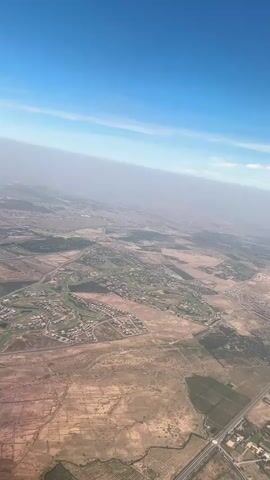 The video depicts an aerial view of a town, taken from a high altitude on a clear day. The camera is positioned looking down at a landscape featuring a mixture of residential areas with clustered houses, a golf course with greenery, and stretches of dry, arid land. Roads and highways cut through the scenery. In the distance, a large city extends into the horizon. A slight haze obscures the farthest reaches of the urban area. The sky is a gradient of light blue near the top of the frame.