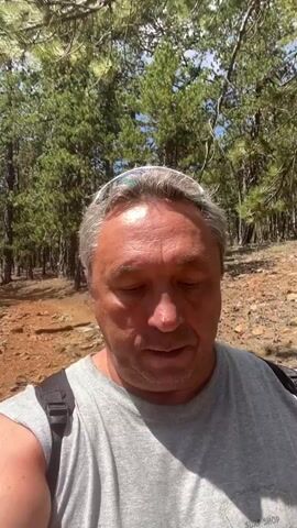 A middle-aged man with graying hair and a grey t-shirt is walking through a forest. He is looking down at the ground as he walks. The trees are tall and thin, and the ground is covered in rocks and dry leaves. The sun is shining through the trees, casting shadows on the ground. He appears to be wearing sunglasses on his head.