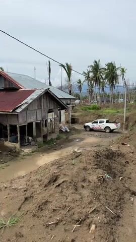 The video opens with a view of a rural or semi-rural area with several houses. One house in the foreground on the left appears dilapidated, with a tin roof and walls made of bamboo and wood. The ground in front of it is muddy, suggesting recent rain or flooding. Further back, more houses are visible, some with tin roofs and concrete structures. A white pickup truck with some writing on its side is parked on a dirt road between the houses. The landscape is dotted with palm trees, and in the distance, there are hills or mountains under a cloudy sky. The overall impression is one of poverty or a recent natural disaster, given the condition of some of the buildings and the muddy terrain.

The second part of the video focuses on a slightly different angle, showing a pile of dirt or debris in the foreground. Two men are standing on this pile, looking towards the houses. One man is wearing a light blue shirt and jeans, and the other is wearing a pink shirt and dark pants. Their interaction is not clear, but they appear to be observing the surroundings. The background remains similar, with houses and palm trees under a cloudy sky. The presence of the men and the truck might suggest some form of aid or assessment after a disaster.