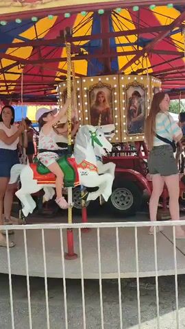 The video captures a carousel ride at what appears to be a fair or carnival. The carousel is adorned with a colorful, striped canopy and features several carousel horses. People of various ages are riding the carousel. In the foreground, a woman in a brown dress walks alongside the carousel while a child on a white horse is visible. Further back, a woman in a gray tank top and black shorts stands on the carousel with a young child in an orange shirt. Another woman in a purple top and blue shorts is riding a white horse. A man in a blue shirt and a child in a blue shirt are also on the carousel. The carousel is enclosed by a white fence.