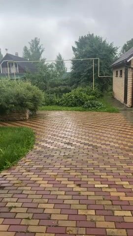 A slightly blurry video shows a red and yellow brick pathway leading into a yard. The pathway is wet, suggesting it's raining or has recently rained. To the left of the pathway is a patch of green grass, and behind that, a lush green bush. Further into the yard, there are trees and two houses. One house is visible in the background to the left, and another structure, possibly a shed or garage, is on the right side of the frame. A yellow pipe runs horizontally in front of the trees and houses. The sky appears overcast. The camera is panning slightly to the right as it moves forward. There are no people visible in the video.