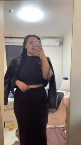 A woman is in front of a mirror, taking a selfie with her smartphone. She is wearing a matching brown crop top and maxi skirt. She also has a brown leather jacket draped over her shoulders. The woman is posing and blowing kisses at the camera. The room has a bed with a white comforter, a window with drawn curtains, and a ceiling light. Some clothes are scattered on the floor near the mirror.