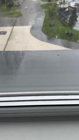 A video shows a view from a window looking out at an outdoor courtyard with buildings in the background. It is raining, with visible raindrops on the windowpane and falling outside. The courtyard has paved walkways, green grassy areas with trees, and a playground with equipment. The buildings are modern apartment complexes. The overall environment appears to be overcast and wet due to the rain. There are no people visible in the video.