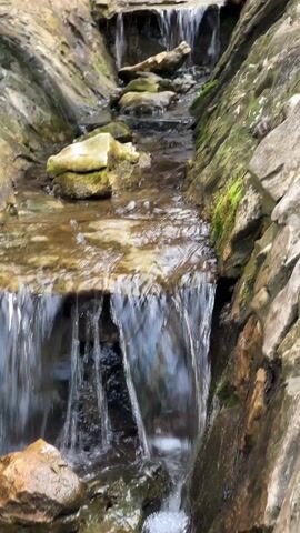 The video shows a small man-made waterfall cascading through a narrow rock gully. The water flows over flat rocks forming small steps, creating multiple miniature waterfalls. The rocks on either side of the stream are covered in patches of moss. A dark wooden structure is visible above the waterfall, obscuring the view of the upper stream.