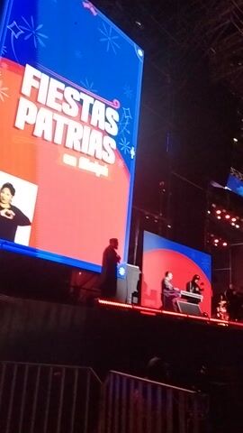 A large, vertically oriented projection screen dominates the frame, displaying a festive graphic for "FIESTAS PATRIAS en Maipú" (Patriotic Holidays in Maipú). The screen features a split design with a blue left side and a red right side, both adorned with white stylized snowflakes or star patterns along their borders.

On the lower right portion of this main screen, a young man with dark hair, wearing a dark long-sleeved shirt, is shown from the chest up, smiling broadly and gesturing with both hands.

In the upper right background, a smaller, elevated stage or screen is visible, also illuminated with red and blue lights. Two dark figures are present on this platform; one appears to be standing behind a console or equipment, and the other is seated. Red stage lights are visible along the side edge of this upper platform.

The surrounding environment is dark, suggesting an evening or nighttime event, likely taking place in a large venue such as an arena or an outdoor concert setting. The audio includes enthusiastic cheering and applause, indicating a lively atmosphere and an engaged audience at this celebration in Maipú, Chile.