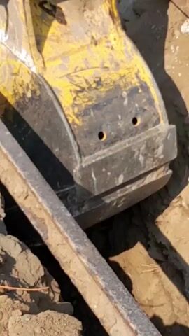 A close-up shot shows a yellow excavator bucket digging into the ground. The bucket's teeth are visible as it scrapes against dirt and debris. The surrounding area is a construction site with loose soil, rocks, and some fragmented building materials. The video captures the dynamic action of the excavator's bucket as it moves, excavating the earth.