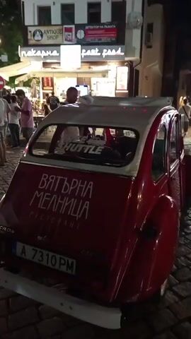 The video shows a narrow cobblestone street at night, lined with buildings that appear to be shops and restaurants. Several people are walking and standing around, some in small groups. In the foreground, the back of a red vintage car is visible, with text on its trunk and a sticker on the rear window. The car has a license plate that reads "A 7310 PM". A sign on the trunk says "ВЯТЪРНА МЕЛНИЦА РЕСТОРАНТ". In the background, shops with illuminated signs are visible, including one labeled "Antique Shop" and another that seems to be a bakery. The overall atmosphere is that of a lively evening in a European town or city.