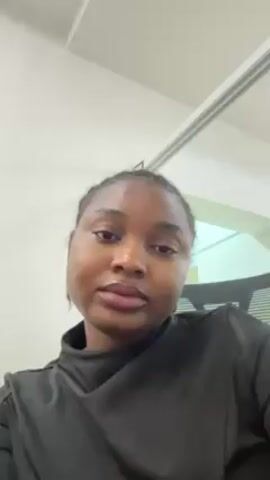 A woman with brown skin and dark hair pulled back from her face is centered in the frame. She is wearing a black long-sleeved top. She smiles at the camera. The background consists of a white wall, a section of a fluorescent light fixture, and a glimpse of an office chair.