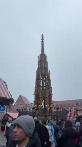 The video shows a tall, ornate fountain, surrounded by a crowd of people. The fountain has a complex structure with multiple levels and statues. It is predominantly dark in color with gold accents. The sky is overcast and gray. The people are wearing winter clothing, suggesting it is a cold day. The background includes buildings with red-tiled roofs and market stalls with red and white striped awnings. There is also a street sign visible. The camera slowly zooms in on the fountain.