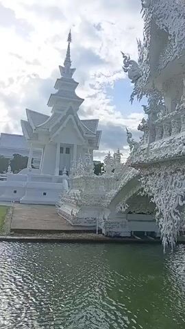 A video clip shows the exterior of a white, ornate building, possibly a temple, with intricate carvings. The building is situated next to a body of water, reflecting the sky which is partly cloudy. The camera pans slowly across the structure, highlighting its detailed architecture. The overall scene is serene and visually striking due to the white color and the detailed ornamentation of the building.