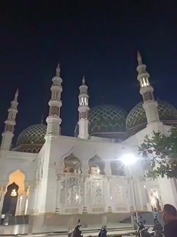 The video is a nighttime shot of a mosque with several minarets and green domes. In the sky, there are what appear to be lights or lanterns in a formation. The mosque is lit up, and there are some trees in the foreground. A few people are visible in the lower part of the frame. The sky is dark, indicating it is nighttime.