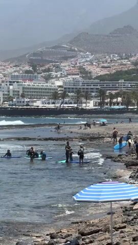 The video shows a rocky beach with people on paddleboards in the shallow water. In the background, a resort town is visible with buildings and palm trees. The sky is hazy and the mountains in the distance are obscured by haze. A blue and white striped umbrella is in the foreground. Some people appear to be taking a lesson as they are gathered with an instructor.