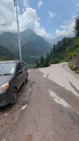 A stationary black car is parked on the left side of a narrow, unpaved road. The road curves to the right, leading into a valley surrounded by lush green mountains. The sky is partly cloudy with sunlight filtering through. Power lines are visible overhead. The car is facing away from the camera. The road appears wet in places.
