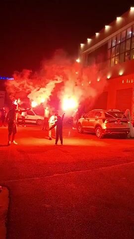 The video captures a nighttime scene illuminated by bright red flares. Several individuals are present, some holding flares that emit dense smoke. Two people in the foreground appear to be the focus, one with arms raised. Cars are visible in the background, including a silver SUV and a white van. The setting seems to be an outdoor area, possibly a parking lot or street, with a modern building featuring illuminated windows in the background. The overall atmosphere is one of excitement or celebration.