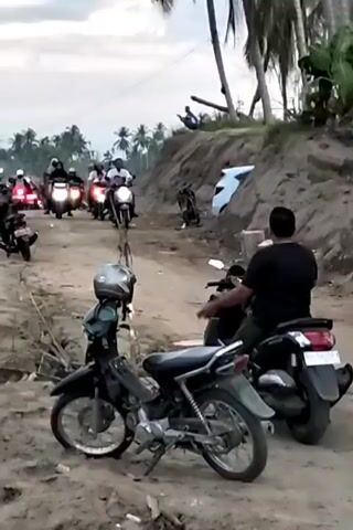 The video shows a muddy road lined with parked motorcycles and people on motorcycles. On the right of the road is an earthen bank. Some people stand on top of the bank, looking towards the water. The camera pans to show the water. On the bank is a dilapidated boardwalk leading to a floating platform, where a small boat is moored.  Palm trees stand in the background.  The sky is overcast.