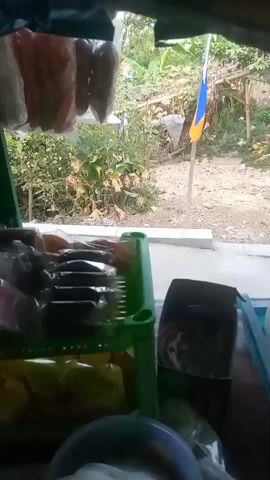 This video shows a close-up view of goods for sale, likely from a street stall or small shop. The items are mostly packaged snacks, including what appear to be sweets and chips. The goods are displayed in plastic bags and containers, with a green plastic tray holding several rows of individually wrapped items in the foreground. Beyond the immediate display, a view of an outdoor scene is visible through an opening. This outdoor scene includes greenery, a road, a house with a balcony, and several flags or banners. The lighting suggests it is daytime. The camera appears to be positioned inside looking outwards.