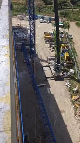 This video captures an aerial view of a construction site in Mirnovskoye rural settlement, Ukraine. The primary focus is on a large blue tower crane, with its boom extending diagonally across the frame. A smaller, green crane is also visible in the background. Several workers are present on the ground, dressed in work attire. Construction materials, including what appear to be concrete blocks and yellow metal beams, are stacked around the site. The scene is outdoors, under what seems to be bright daylight, suggesting a clear weather day. The overall atmosphere is one of ongoing construction and industrial activity. No visible text is present in the video.