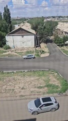 The video shows a street scene with several cars and buildings. In the foreground, a white SUV with a sunroof is parked on a paved road. Behind the SUV, a gray sedan is driving on the same road, which appears to be in a rural or suburban area. The buildings visible in the background are mostly single-story houses with pitched roofs, some of which appear to be in disrepair. There are trees and dry vegetation throughout the scene, and the sky is partly cloudy. The video is shot from a high vantage point, looking down on the street.