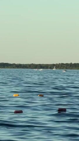 A white speedboat with a black outboard motor races across a body of water. The boat is leaving a white wake behind it. In the foreground of the image are several buoys, some yellow and some red, floating on the water. In the background, a tree line is visible along the horizon under a clear, pale blue sky. The camera is positioned low on the water, giving the impression that it is also on a boat.