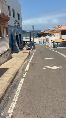 The video displays a bright, sunny daytime scene of a quiet street in a coastal town, likely Pájara, Spain. The asphalt road, marked with white lines and two painted white pedestrian figures, slopes gently downwards towards the sea, which is visible in the distance under a clear blue sky.

On the left side of the street is a white two-story building with a vibrant blue lower section, featuring a mural-like design. This building houses a "RESTAURANTE CAFE BAR," with tables and chairs set out under an awning. A menu board stands on the tiled sidewalk in front of the restaurant.

On the right side of the street, there are other white buildings with blue accents, typical of coastal architecture. A street lamp stands near the restaurant, and another is visible further down the street. Rocky cliffs and a rugged coastline are seen in the background, framing the view of the ocean. The overall atmosphere is peaceful and serene, with no people actively moving in the frame.