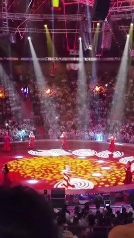 A wide shot of a circus performance in progress. The stage, illuminated by spotlights and patterned lighting, features several performers in red dresses dancing in a circle. In the background, a large audience fills the stadium seating. High above the stage, a performer is suspended from the ceiling by a wire, performing an aerial maneuver. The atmosphere is lively, with colorful lighting and a sense of spectacle.
