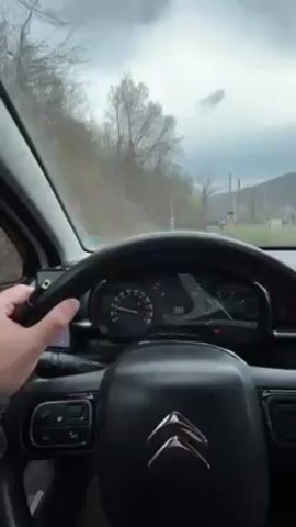 The view from inside a car is shown in this video. The car is approaching a railway crossing. The car's speedometer indicates decreasing speed as the driver slows down. The driver's hand is visible on the steering wheel. Beyond the railway crossing, trees and hills are visible under a cloudy sky. The car's dashboard and interior are visible, including the rearview mirror with a small ornament hanging from it. The time on the car's display reads 12:33. Music is playing in the background.