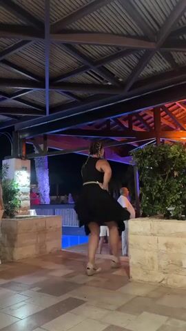 A woman in a black dress is dancing at night outdoors. She is wearing white wedge sandals and her hair is in a braid. The dress is short and flares out as she spins. She is dancing in front of a pool and there are other people in the background, some standing and watching, and others appearing to be dining at tables. The area is covered by a wooden roof with lights that cast an orange glow. There is also a large green bush to the right of the woman and stone walls in the foreground.
