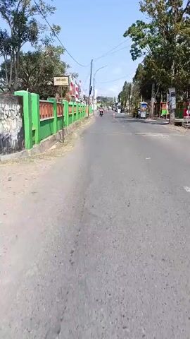 The video shows a perspective from a vehicle, likely a motorcycle, traveling down a paved road. The road is lined with green fences and trees on both sides. Indonesian flags are visible, suggesting a celebration or patriotic occasion. There are several small stalls or carts on the side of the road, and a few people are seen walking or sitting. The sky is clear and blue, indicating a sunny day. The camera pans and moves forward, capturing the scenery as the vehicle progresses.