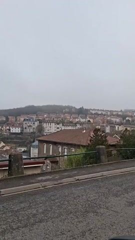 The video shows a view of a town on a foggy day. The houses are mostly light-colored with red tile roofs. The camera is positioned on a street overlooking the town, with a concrete barrier and some bushes in the foreground. The sky is overcast and grey. The road is asphalt. Some music plays in the background.