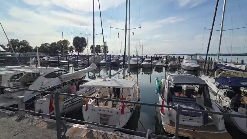 This is a daytime exterior shot of a marina filled with various boats, mostly sailboats and yachts, docked side-by-side. The water is calm and reflects the boats and the sky, which is partly cloudy with the sun visible. A walkway with a metal railing runs along the edge of the marina. Trees and buildings are visible in the background on the shore. One person can be seen on one of the boats, appearing to be relaxing or preparing to depart. The boats are white and a few have flags displayed, including German flags. The overall atmosphere is peaceful and sunny.