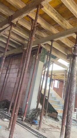 The video captures a close-up view of a construction site in Petrovo, Bulgaria, focusing on the scaffolding and unfinished structure of a building. The scene is characterized by numerous rusty metal support poles (shoring jacks) holding up a wooden ceiling structure made of planks.

In the foreground, a person's leg and foot are visible, wearing a grey athletic shoe with lime green accents and white socks that have the word "FITNESS" printed on them. This suggests the person is taking a break from manual labor or is observing the construction. A black backpack is seen hanging on one of the support poles in the midground.

The background reveals more of the construction site, including brick walls, concrete elements, and stairs leading upwards, indicating the building is in its early stages of construction. The lighting suggests it is daytime, with natural light filtering through gaps in the structure. The overall impression is one of ongoing construction work, with temporary supports in place for a concrete pour or similar structural activity. The presence of the person in the foreground with their foot up implies a moment of rest amidst the work. There is no visible text that provides further context or information.