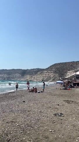 The video shows a beach scene. There are people on the beach and in the water. The beach is covered in pebbles. There are cliffs in the background. The sky is clear and blue. A lounge chair is visible in the foreground.