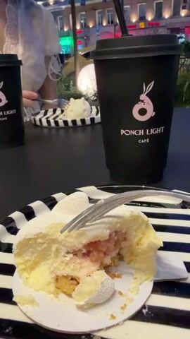 The video shows a close-up of a dessert with a bite taken out of it. The dessert is on a black and white striped plate. Next to it is a black cup with a pink bunny logo and the text "PONCH LIGHT CAFÉ". In the background, a person is visible, partially out of focus, holding a fork and eating. The setting appears to be an outdoor cafe or restaurant with buildings and lights in the background.