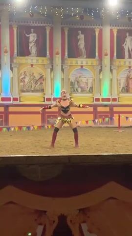 A woman in a gladiator costume dances in an arena. The costume includes a black and gold top, a gold skirt with a red fringe, gold armbands, and red knee-high boots. Her hair is styled up and she has a tiara on her head. She is wearing makeup and jewelry. The arena has a sandy floor and a backdrop with Roman-themed murals and columns. Colorful flags are strung across the front of the arena. The lighting is dramatic, with spotlights and colored lights. The woman is moving her arms and legs in a dance routine. She is holding a sword in one hand. Her expression is one of performance and engagement with the audience. The video is shot from a slightly elevated perspective, looking down into the arena. The focus is on the dancer and her performance.