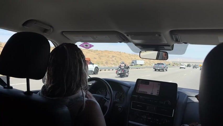 The view from inside a moving car shows the driver, a person with light-colored braided hair and a light-colored top, driving on a highway. The driver is female. There is another person in the passenger seat.  Motorcycles are passing the car. Other cars and trucks are also driving on the highway.  The interior of the car includes a navigation screen, a rearview mirror, and front seat headrests. The outside environment appears to be a desert landscape with hills and clear skies. Wind turbines are visible in the distance. The car is moving at highway speed.  Music is playing in the car.
