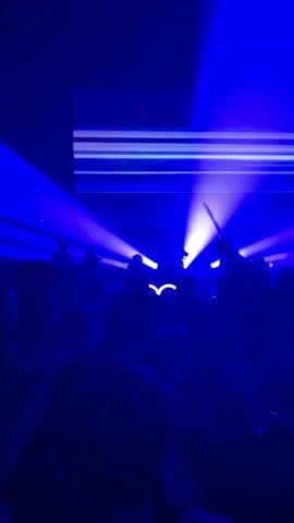This is a vertical video clip from a concert or nightclub. A crowd of people are visible in silhouette, dancing in a dimly lit, blue environment. Stage lighting, consisting of bright blue beams, cuts through the darkness from above, illuminating the space and creating a dramatic atmosphere. A large screen at the top of the frame displays horizontal lines of light, likely part of the visual show. Some audience members are raising their hands in the air, engaged with the music and performance. The overall mood is energetic and celebratory.